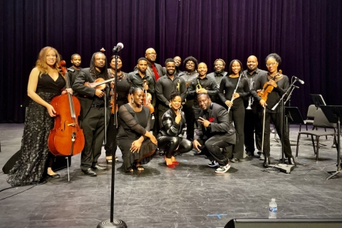 A group of approximately 20 musicians all dressed in black holding classical music instruments