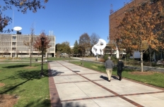 A brick and pavement pathway stretches out away from the viewer. Two men are walking and talking. In the distance you see the undulating bright white ribbons of the Koan sculpture.