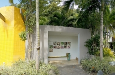 A bright yellow building with an A frame window next to a shaded open room, all surrounded by palm trees